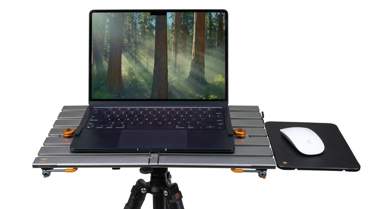 Laptop on a metal platform mounted on a tripod, with a mouse pad and white mouse to the right, displaying a forest wallpaper.