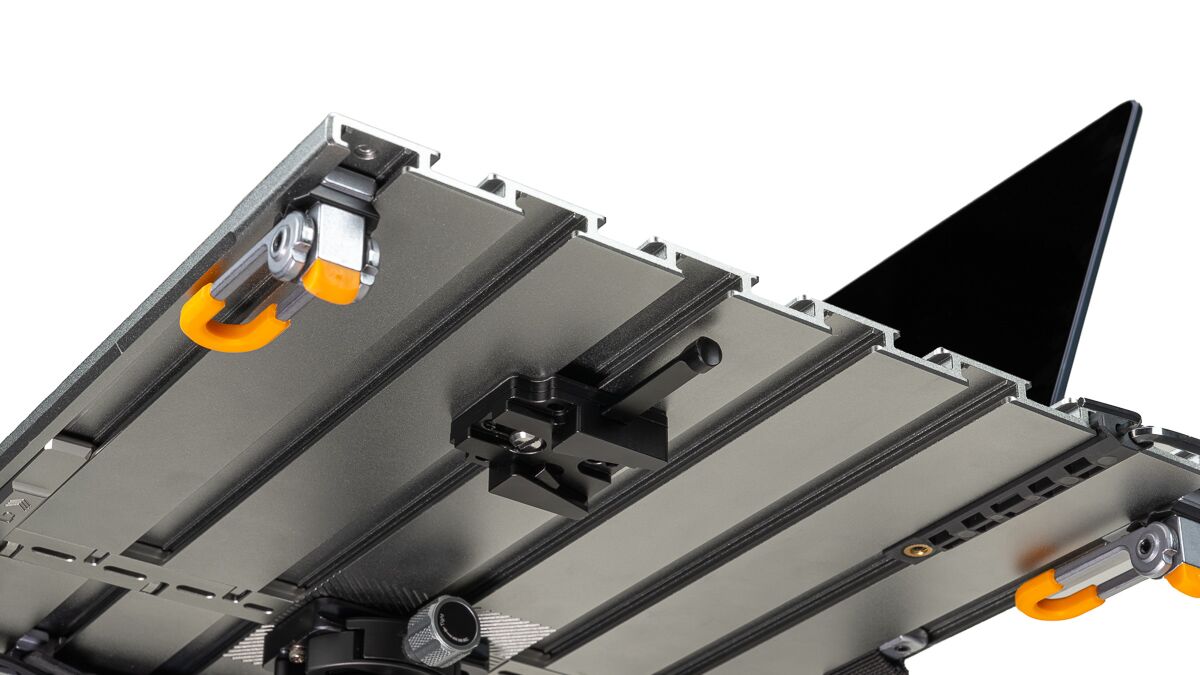 Close-up of a modular camera rig underside with aluminum rails, clamps, and orange locking handles; a laptop is visible in the background.