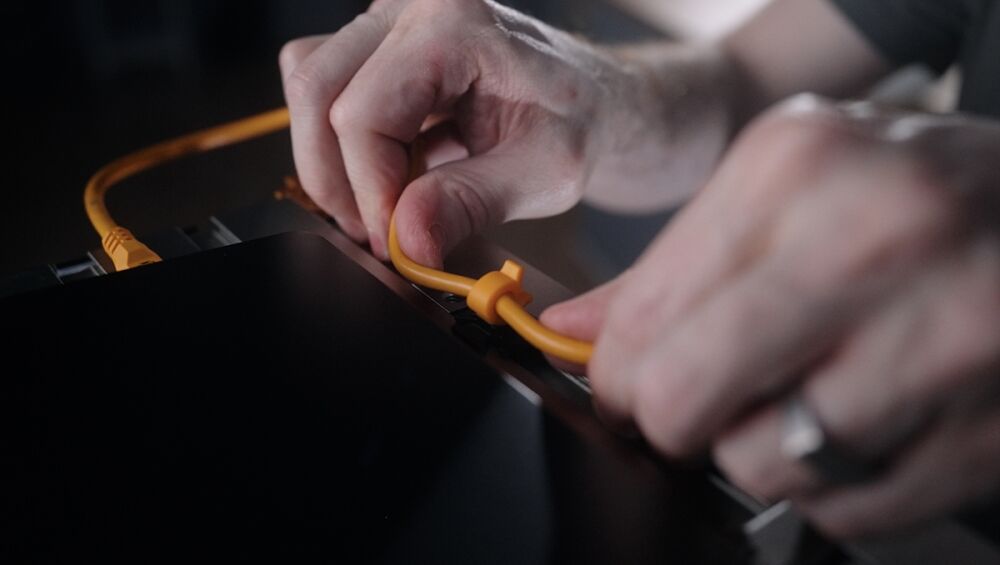 Two hands plug an orange Ethernet cable into a dark electronic device, close-up detail of the connection.",