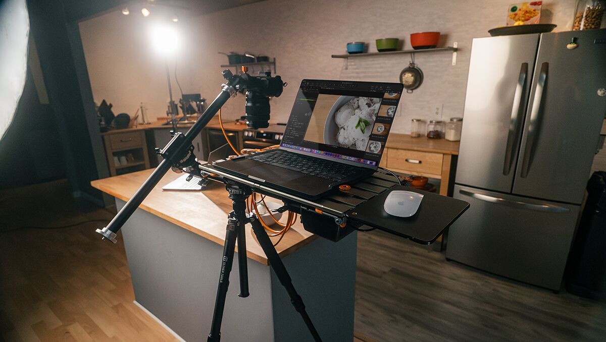 Camera on a tripod with a laptop on a portable desk in a kitchen studio, displaying food photography edits