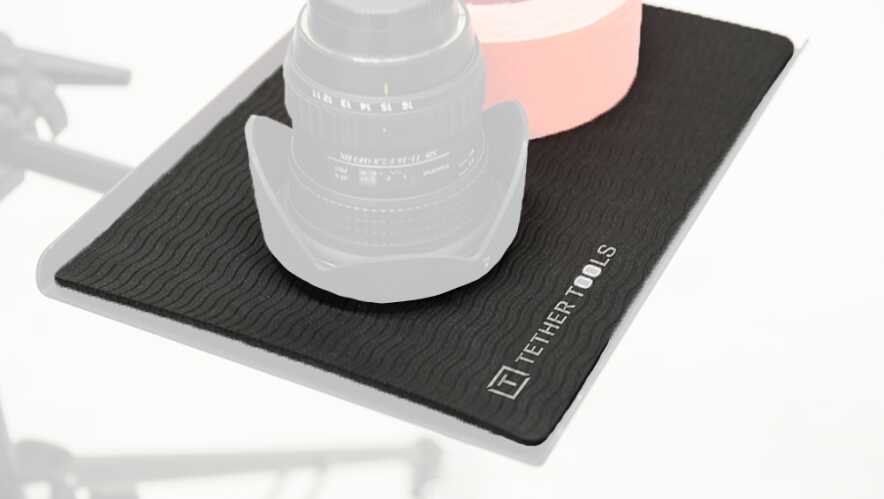 Close-up of a camera lens resting on a black textured anti-slip mat, with a pink roll in the background.