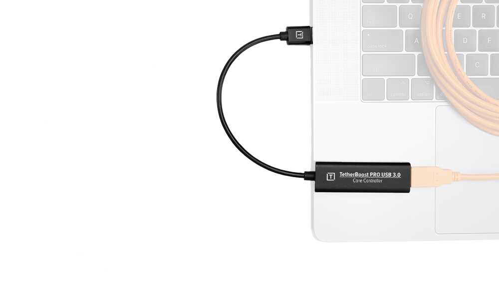 Close-up of a laptop edge with a black USB device labeled 'TetherBoost PRO USB 3.0 Core Controller' connected to a beige USB cable.