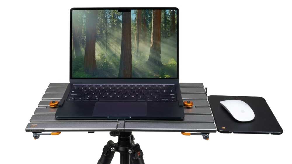Laptop on a metal platform mounted on a tripod, with a mouse pad and white mouse to the right, displaying a forest wallpaper.