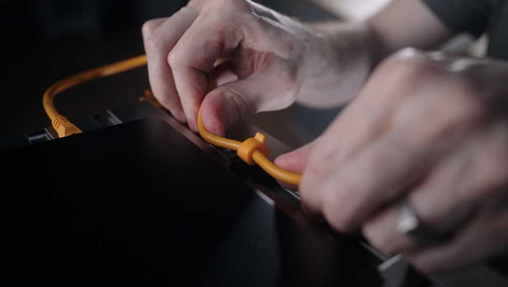 Two hands plug an orange Ethernet cable into a dark electronic device, close-up detail of the connection.",