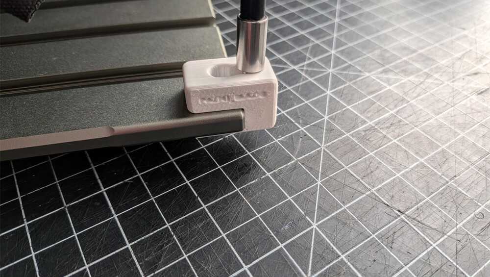 White 3D-printed clamp securing a metal rod to a dark work surface in a workshop setup on a grid cutting mat.