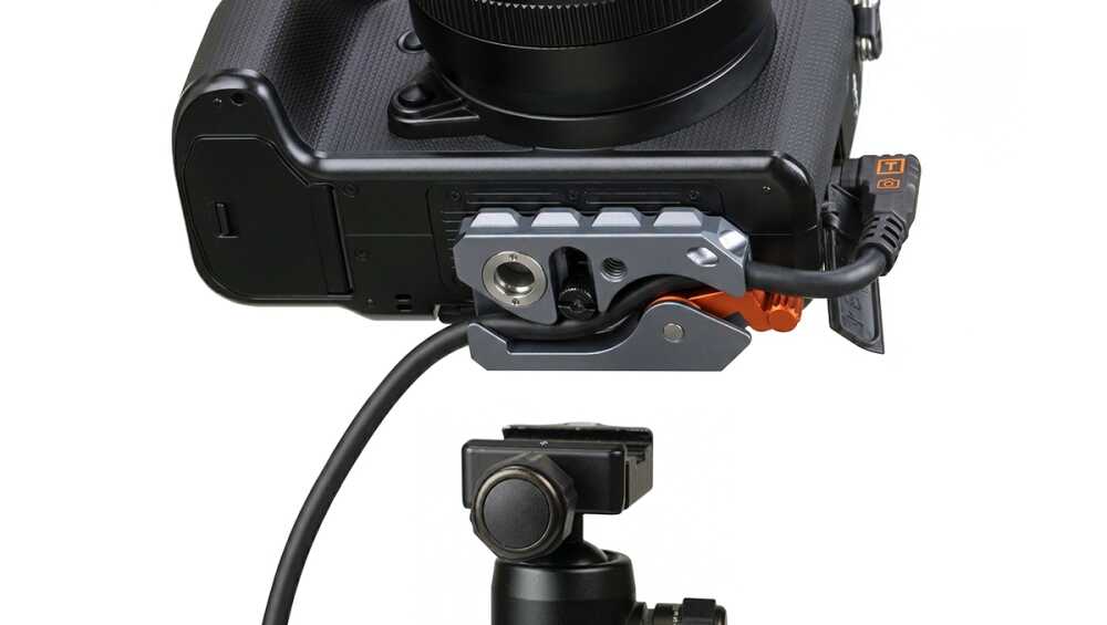 Close-up of a camera's bottom with a modular mounting rig and orange quick-release lever, connected by a cable to an accessory.