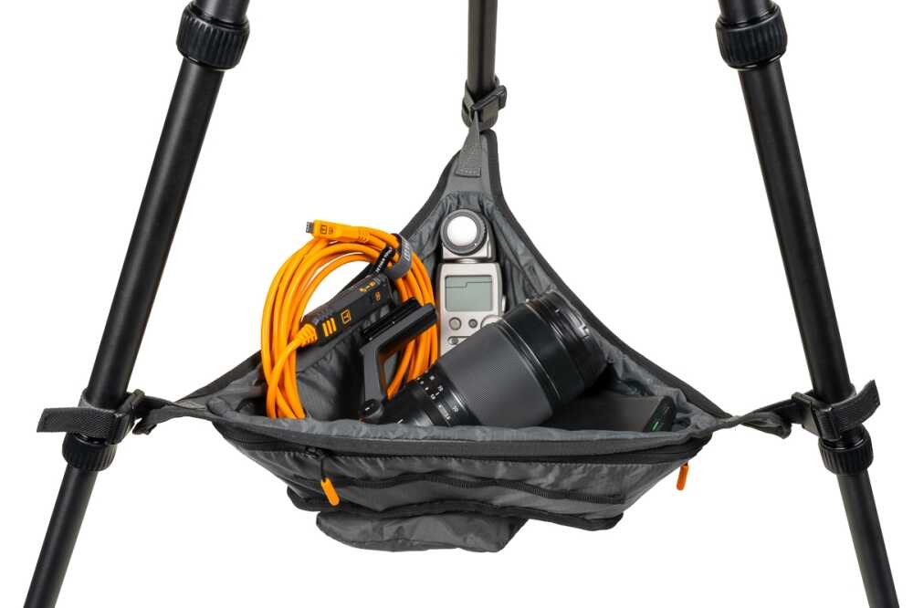 Camera gear stored in a hanging triangle pouch between tripod legs: orange cables, a camera lens, and a small digital device visible inside the bag