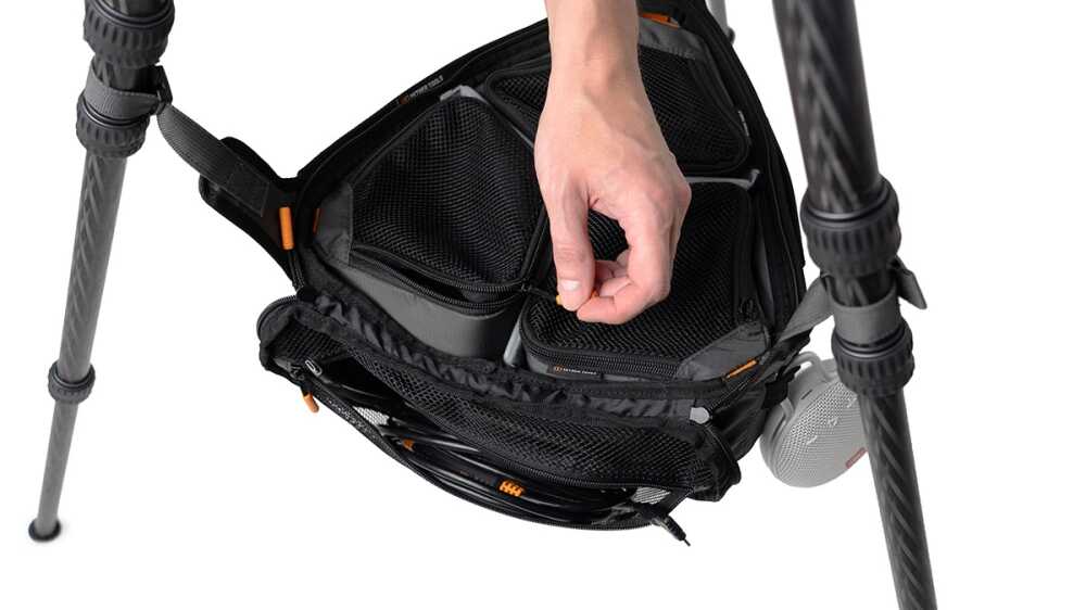 Close-up of a hand reaching into the mesh pocket of a black camera backpack mounted on a tripod.