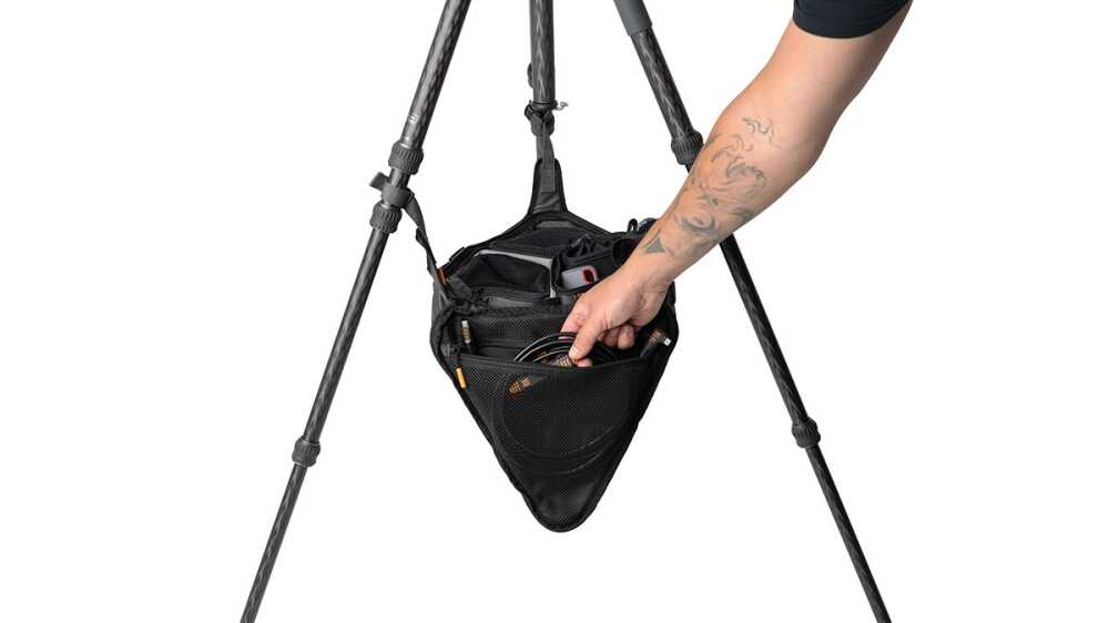 Person with tattooed arm reaching into a black camera gear bag hanging between tripod legs, organizing equipment in a studio setup