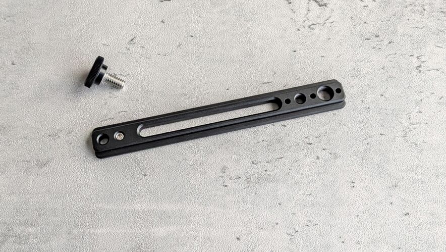 Black slender mounting plate with a long central slot and several holes, shown with a small screw knob nearby in the corner.