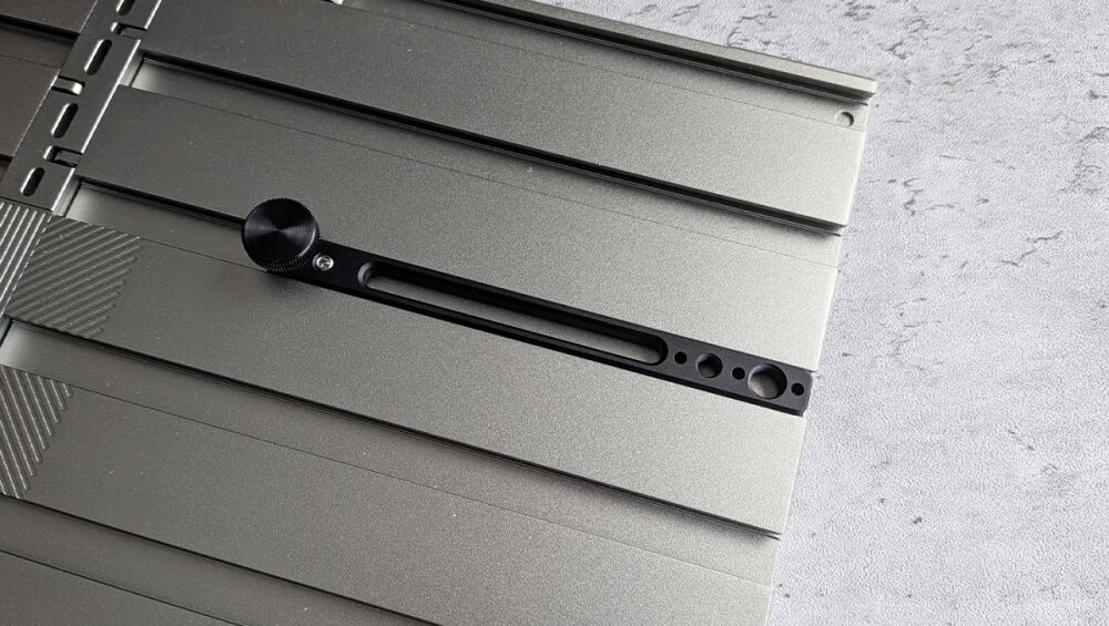 Close-up of a metallic camera slider rail with a black knurled adjustment knob and elongated slot, mounted on a grey surface