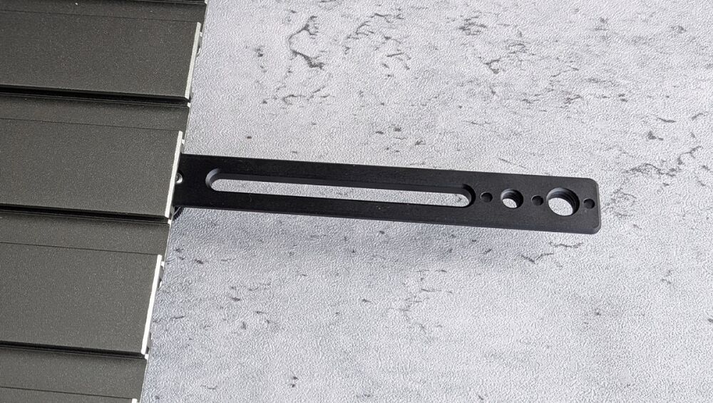 Black metal mounting bracket with a long oval slot and four circular holes, attached to the edge of a dark panel.