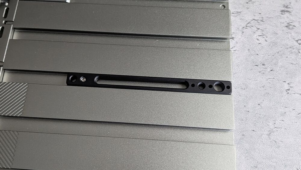 Close-up of a gray metal panel featuring a long black carriage/slot with small circular holes along the edge, likely a mounting or retention area on hardware.