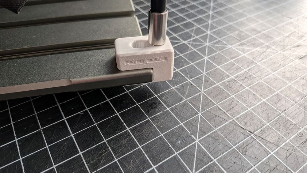 White 3D-printed clamp securing a metal rod to a dark work surface in a workshop setup on a grid cutting mat.