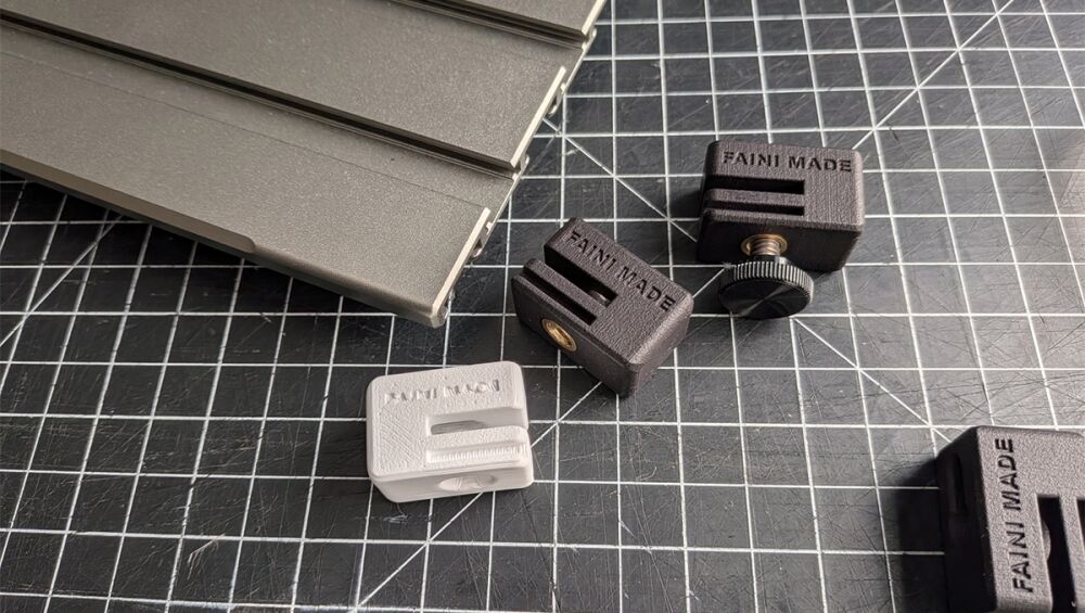 Assorted small plastic clamps on a dark grid cutting mat, with a stack of gray metal pieces to the left.