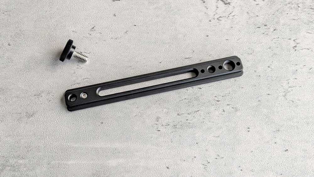 Black slender mounting plate with a long central slot and several holes, shown with a small screw knob nearby in the corner.