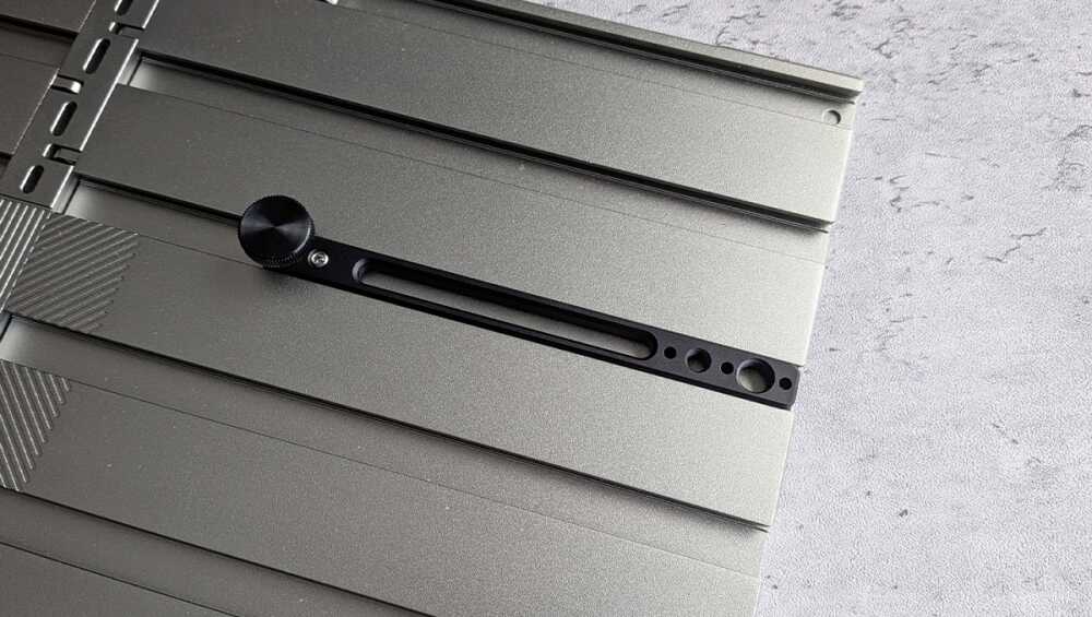 Close-up of a metallic camera slider rail with a black knurled adjustment knob and elongated slot, mounted on a grey surface