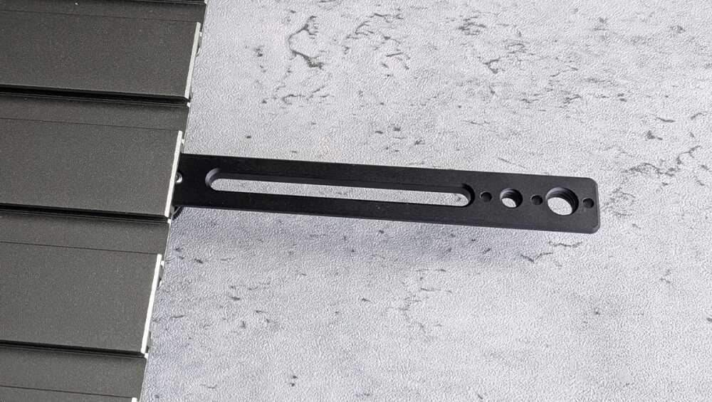 Black metal mounting bracket with a long oval slot and four circular holes, attached to the edge of a dark panel.