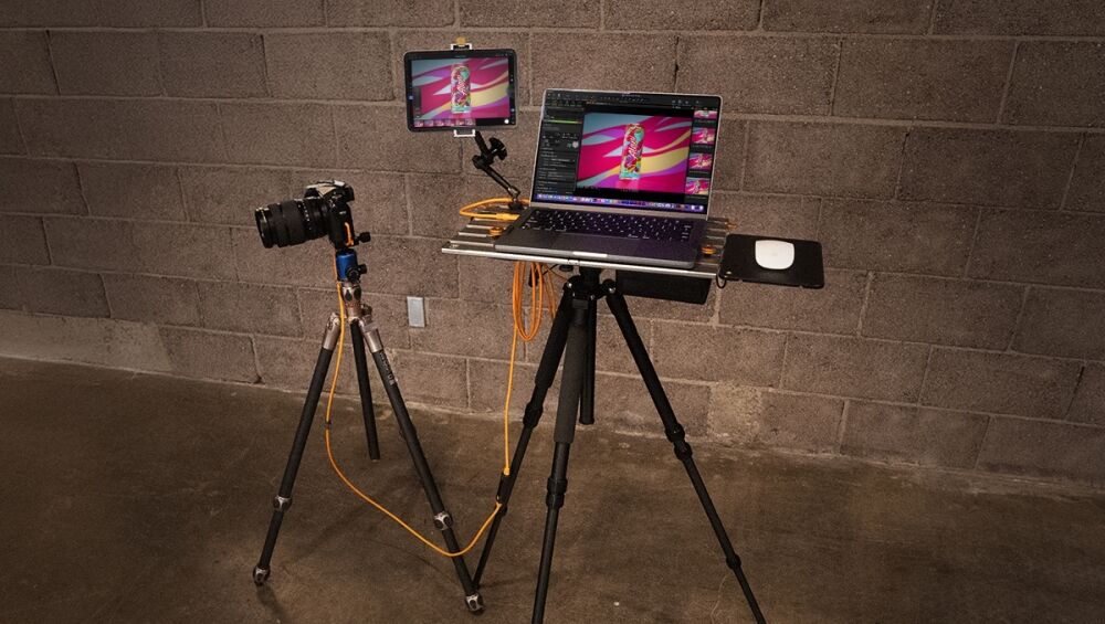 Camera on a tripod connected to a laptop and a tablet displaying a colorful image, with orange cables, against a brick wall.