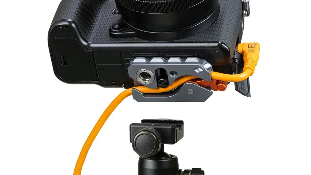 Close-up of a DSLR camera attached to a gray battery grip with an orange power cable plugged into the bottom port, on a tripod head