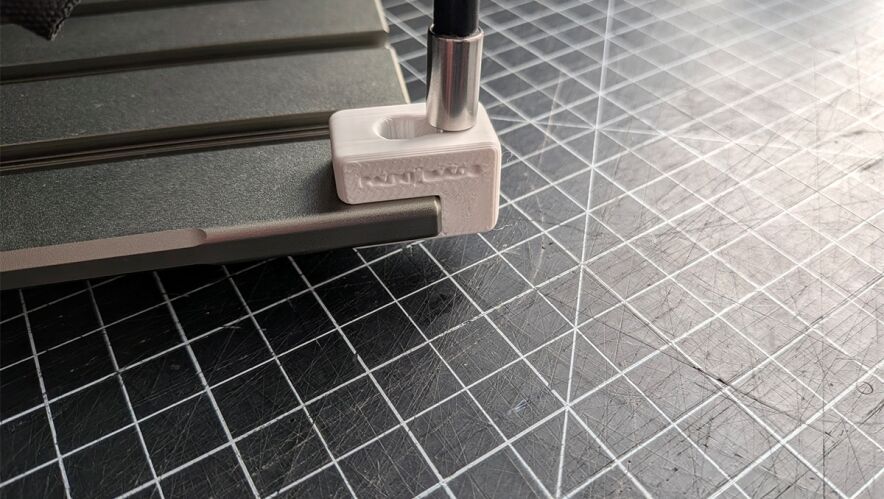 White 3D-printed clamp securing a metal rod to a dark work surface in a workshop setup on a grid cutting mat.